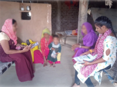 Laxmi, right, speaks with three women during a routine home visit. Two women sit on charpoys while another crouches on the floor beside a small child.